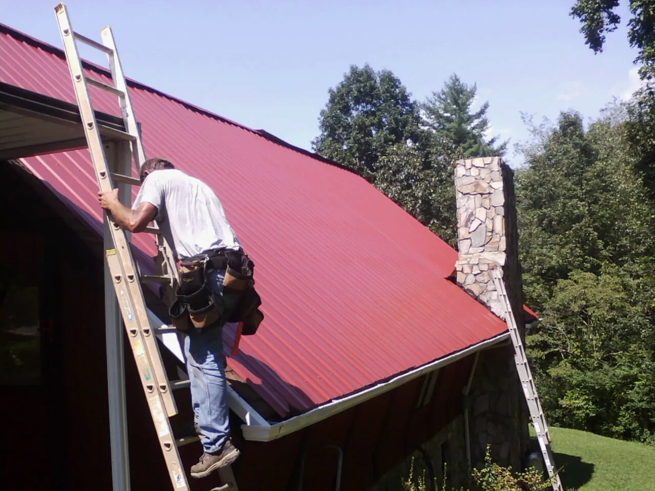 Roofer on a ladder beside a red metal roof and stone chimney — Soffit & Fascia Repair by Roofing Brothers in Todd